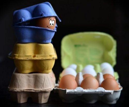 Featured Image Egg Painted With Facial Features Peeks Out Looking Down From 3-Tier Carton Stack Upon An Opened Full Carton Below/Alongside.
